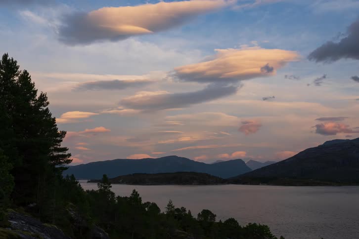 A serene timelapse captures lenticular clouds glowing with colorful sunset light as they drift over a tranquil mountain lake