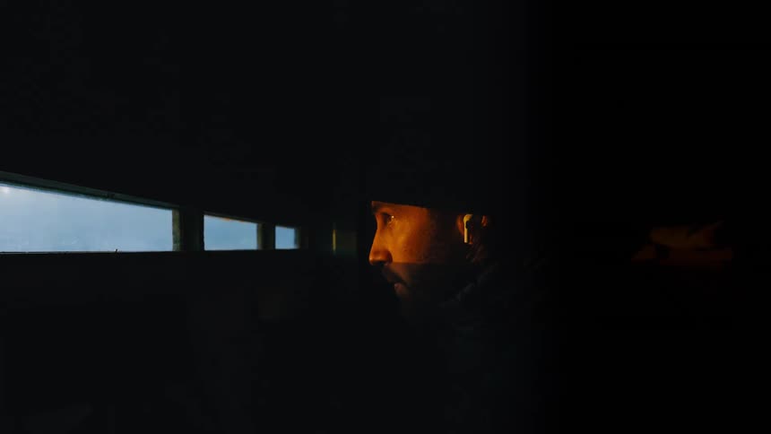 A cinematic close-up shows a contemplative man's face illuminated by a warm, golden light as he looks out a narrow window from a dark room