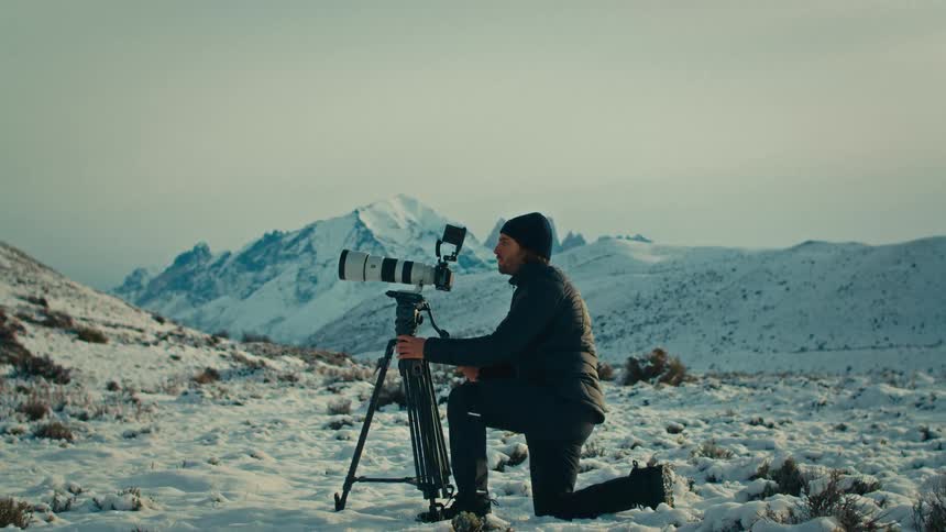 A dedicated photographer kneels in the snow, operating a camera with a telephoto lens on a tripod against a backdrop of majestic, snow-covered mountains