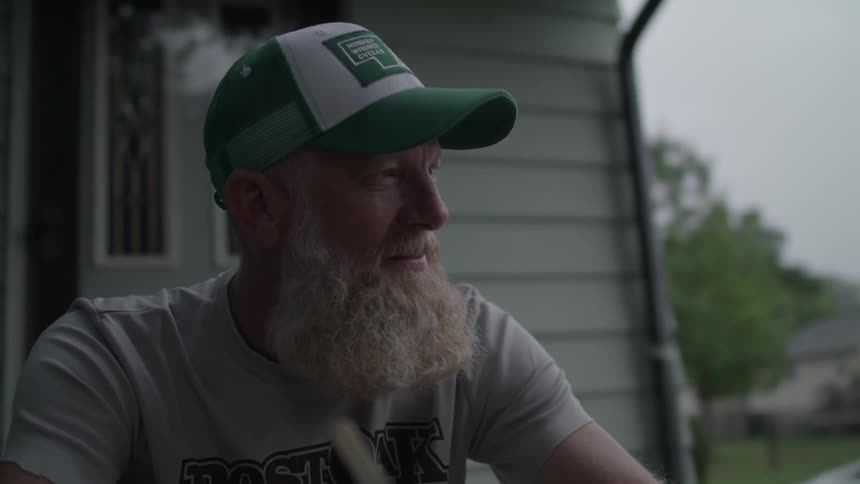 An older man wearing a baseball cap is sitting on his porch