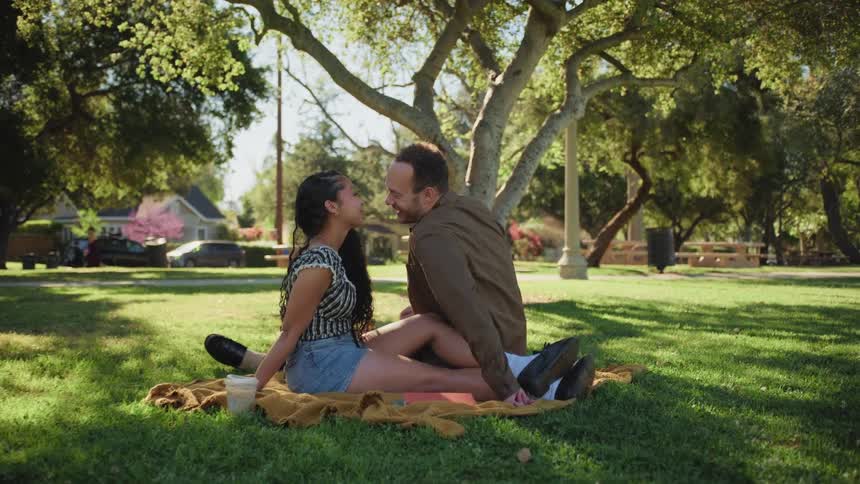 A loving couple shares a romantic moment while sitting on a blanket under a shady tree in a sunny park
