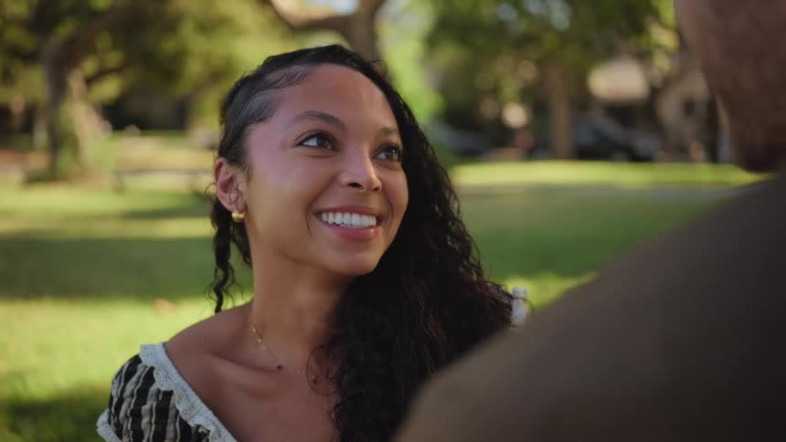 A beautiful young woman with long curly hair smiles lovingly at her partner during a romantic date in a sunny park