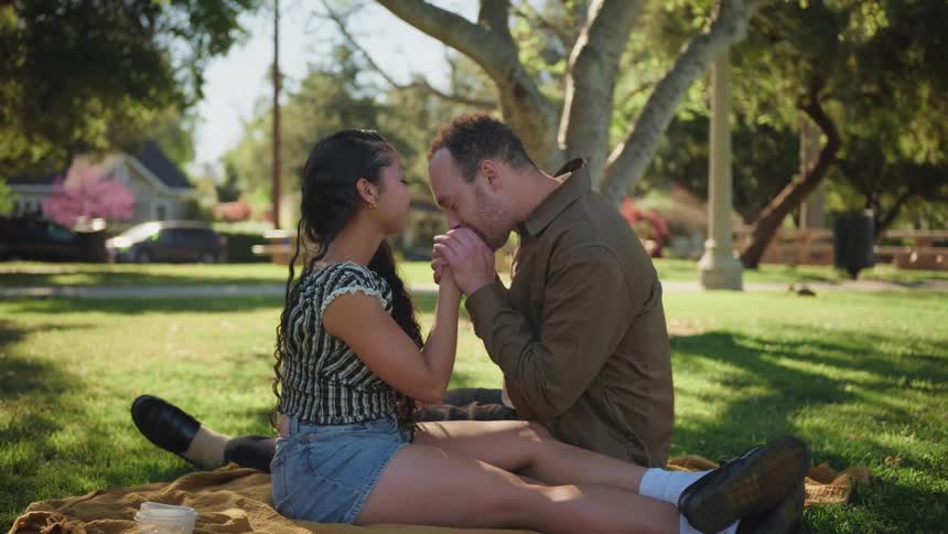 An affectionate couple sits on a blanket in a sunlit park, holding hands and sharing a gentle kiss