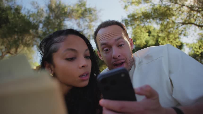 A happy couple relaxes on a blanket in a sunny park, sharing a moment of surprise and laughter while looking at a smartphone