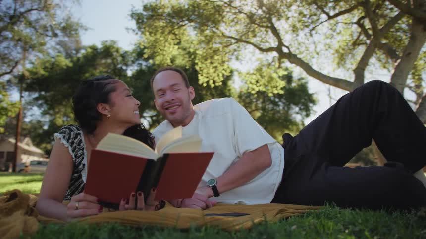 An affectionate couple reads a book together and shares a tender kiss while relaxing on a blanket in a sunny park