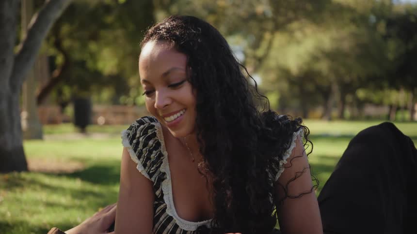 A beautiful young woman with long curly hair smiles while relaxing in a sunny park with her partner