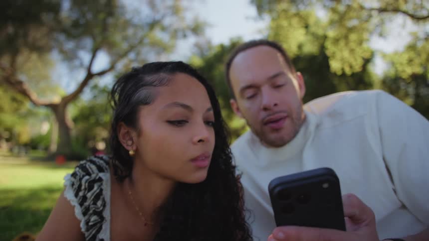 A fast-moving camera pushes in on a young couple relaxing on a blanket in the park, as the man interrupts the woman's reading to show her something on his phone