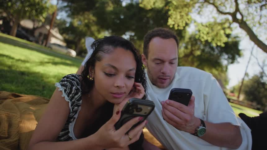 A spinning camera effect reveals a happy young couple lying on a blanket in the park, smiling as they look at their smartphones