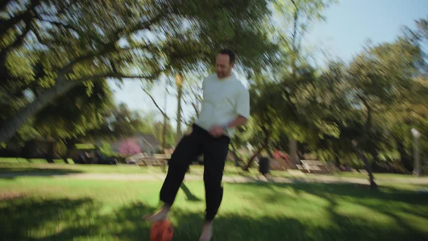Dynamic handheld shot of a barefoot man dribbling a soccer ball through a sunny park