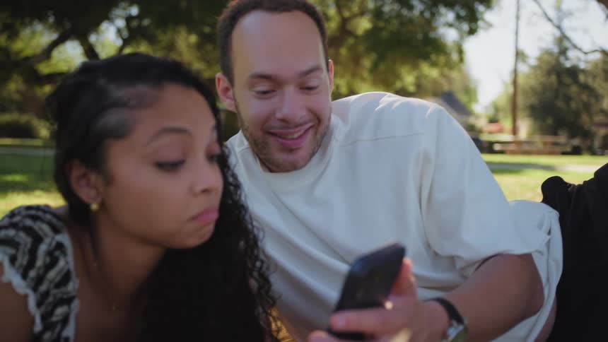 A smiling young man shows his girlfriend something on his smartphone while they relax on a blanket in a park