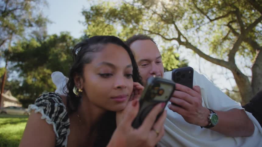 A dynamic dolly-in shot reveals a young couple relaxing on a blanket in a sunny park, both absorbed in their smartphones