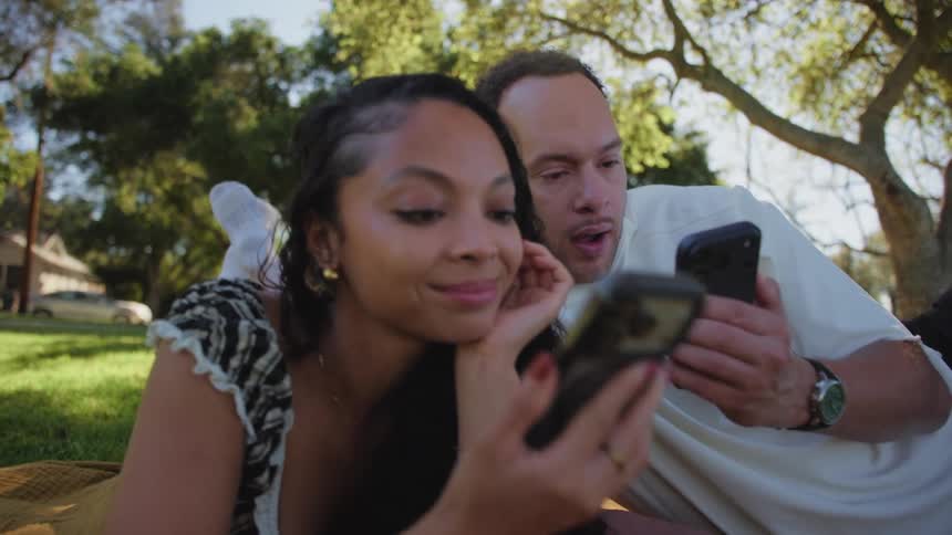 An interracial couple relaxes on a picnic blanket in a sunny park while scrolling on their smartphones together