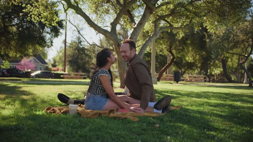 A loving couple shares a kiss and a smile while having a romantic picnic on the grass in a sunny park
