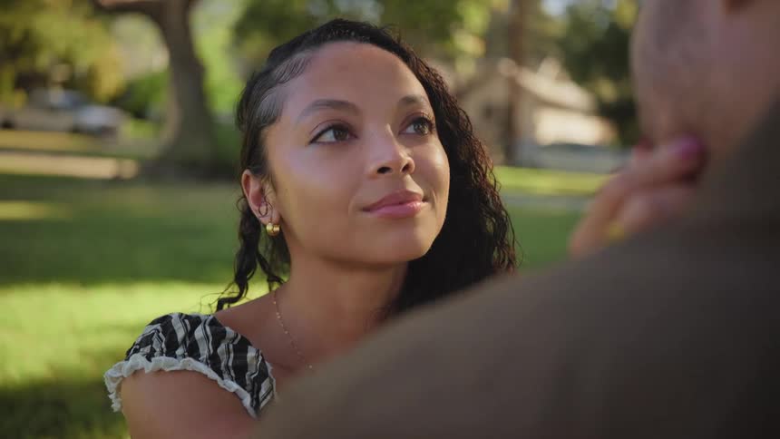 A young woman gazes lovingly into her partner's eyes, her expression softening into a warm smile as she affectionately caresses his face in a sunlit park