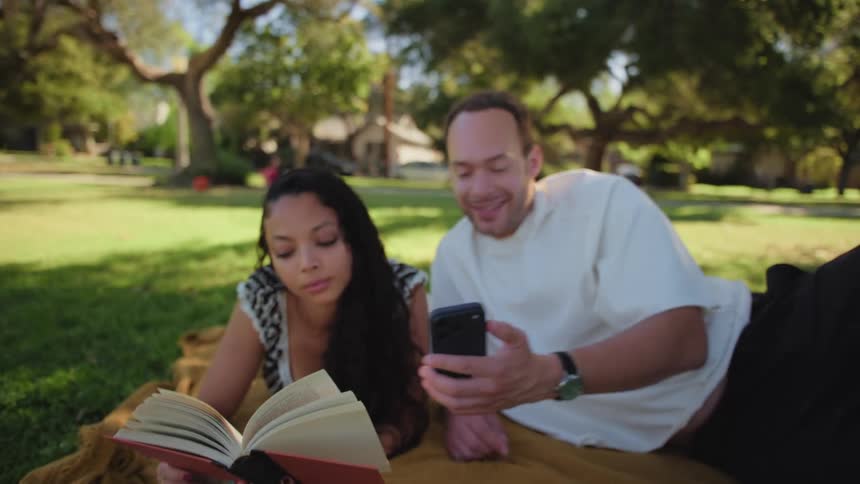 A man shows his girlfriend something on his smartphone while she is reading a book, and they laugh together while relaxing on a blanket in a sunny park