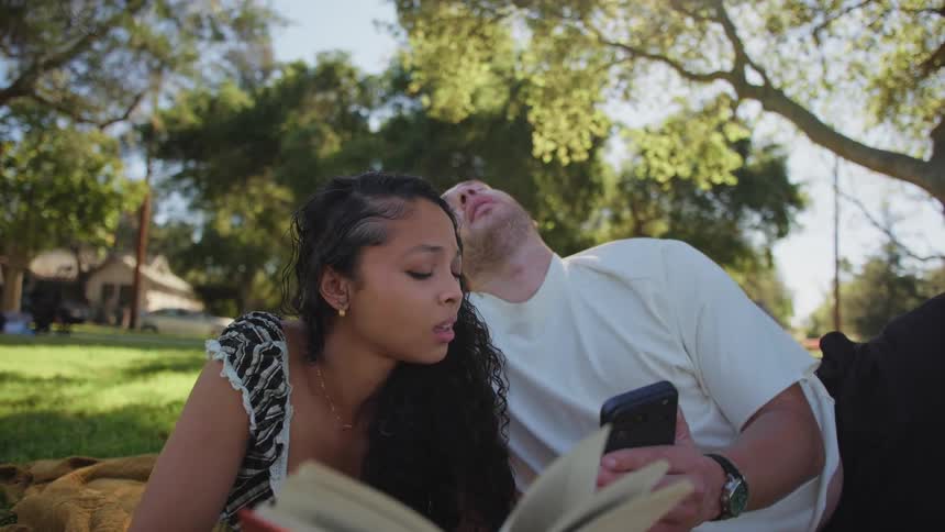 A loving couple relaxes on a blanket in a sunny park, as the woman reads a book and the man shows her something on his phone