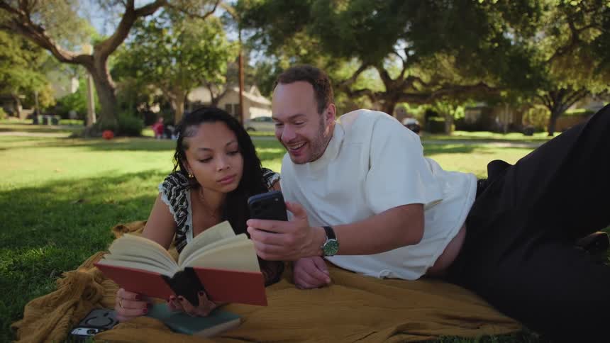 A dynamic spinning shot reveals a happy couple on a park blanket, where the man distracts his partner from her book to share a laugh over his phone