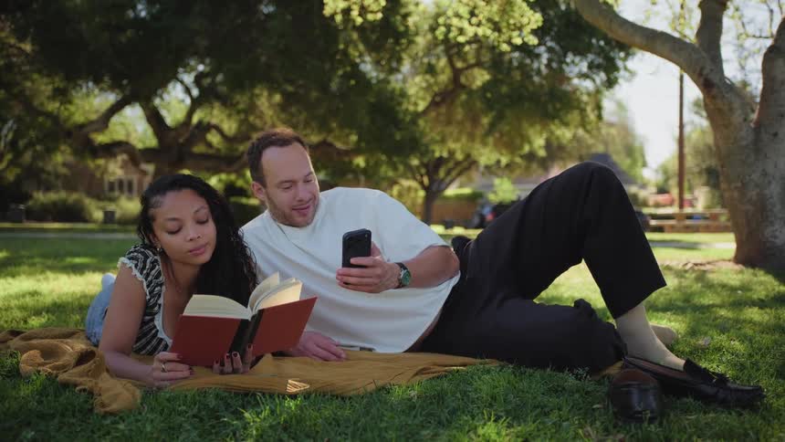 A woman reads a book while her partner uses his phone as they relax together on a blanket in a sun-dappled park