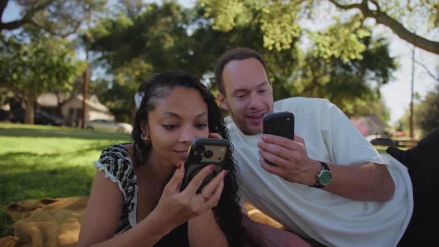 A diverse couple relaxes on a blanket in a park, smiling and laughing as they scroll through their smartphones together on a sunny day