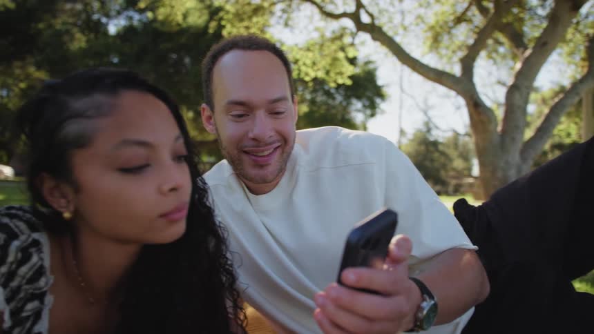 A happy couple relaxes on a blanket in a sunny park, laughing together while looking at a smartphone