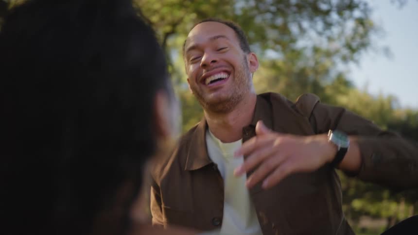 An expressive man sitting in a park enthusiastically tells a story to a friend, bursting into joyful laughter