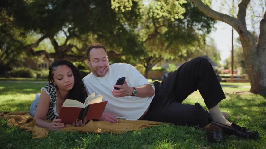 A young couple spends a relaxing afternoon in a sunny park, where a woman reads a book while a man scrolls on his phone and shows her something funny