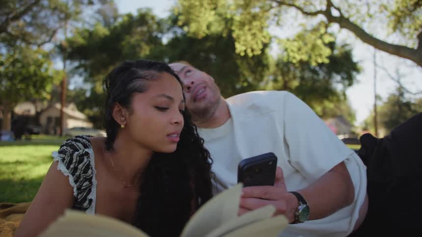 A diverse couple relaxes on a blanket in a sunlit park, reading a book and looking at a smartphone together