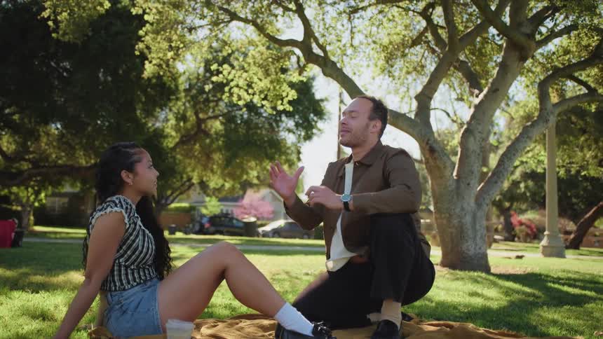 A man animatedly tells a story to a woman as they sit on a blanket during a date in a sunny park
