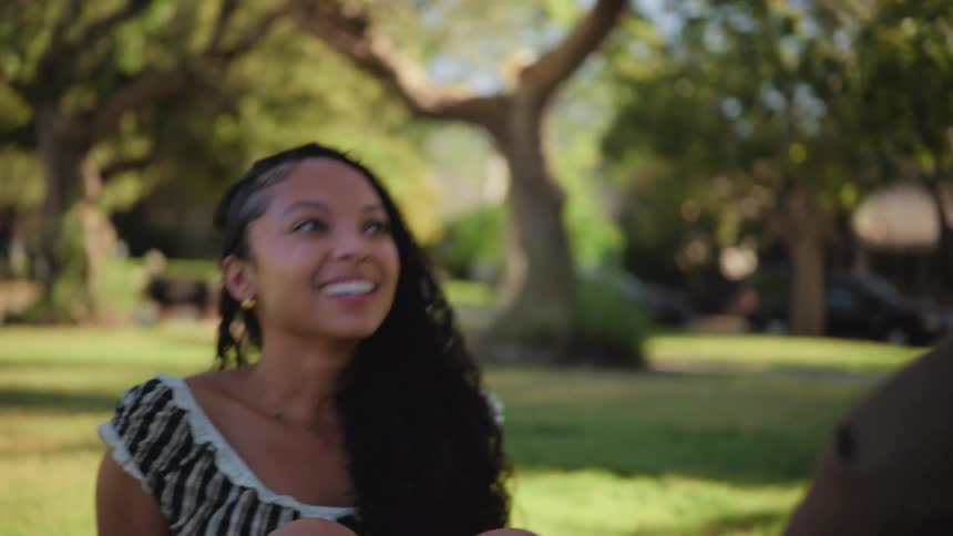 A beautiful young woman smiles and laughs while having a pleasant conversation with her date in a sun-dappled park