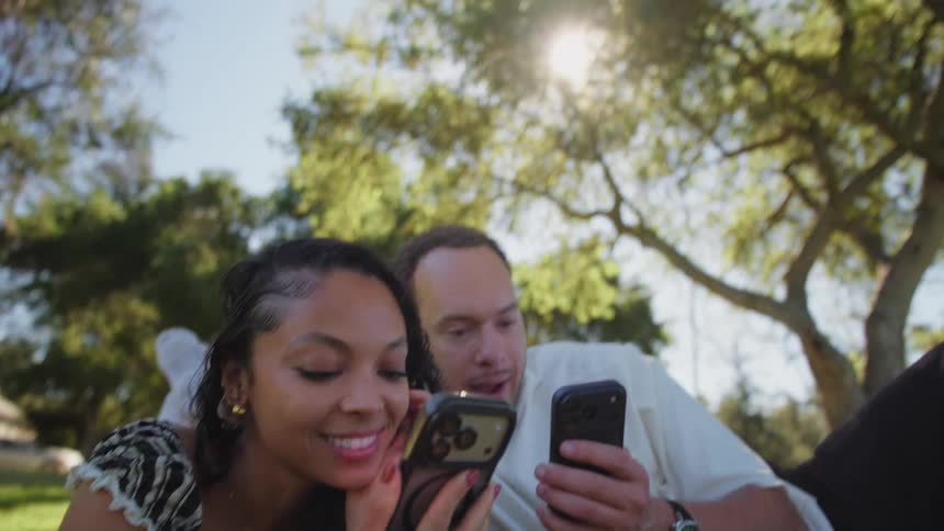 A happy couple lies on a blanket in a park on a sunny day, laughing as they look at their smartphones together