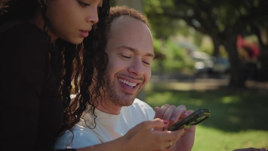 A happy couple enjoys a sunny day in the park, smiling as they look at a smartphone together