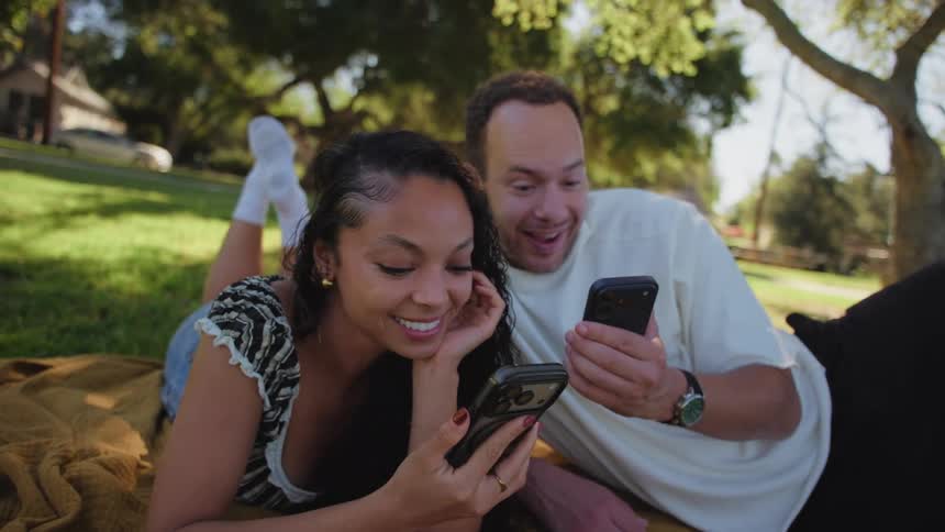 A dizzying spin reveals a happy couple lying on a blanket in a park, smiling together as they look at their phones