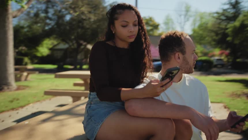 An interracial couple sits disengaged from one another at a park table, both absorbed in their smartphones