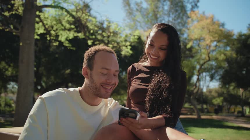 A loving young couple sits closely together in a sunny park, smiling as they look at a smartphone