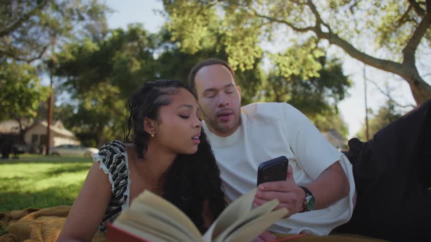 A young couple relaxes together on a blanket in a sunny park, reading a book and looking at a smartphone
