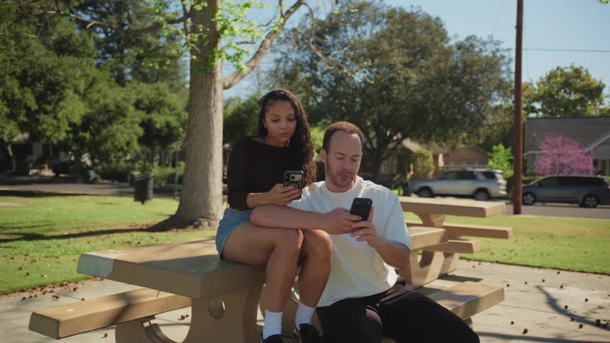 A disconnected couple sits together at a picnic table in a park, each absorbed by their own smartphone
