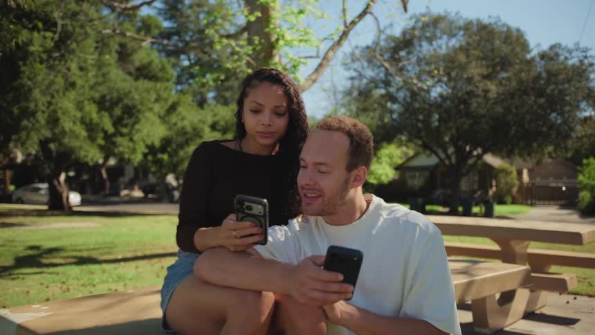 A young couple sits together at a park picnic table, both engrossed in their smartphones on a sunny afternoon