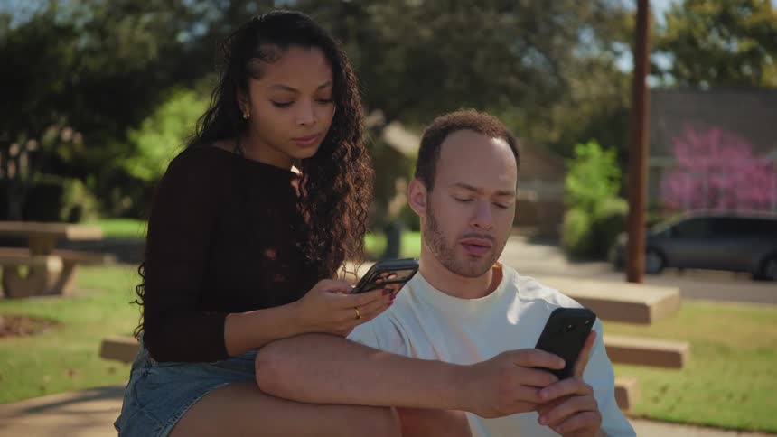 A young couple sits closely together in a park, both absorbed in their smartphones and disconnected from one another