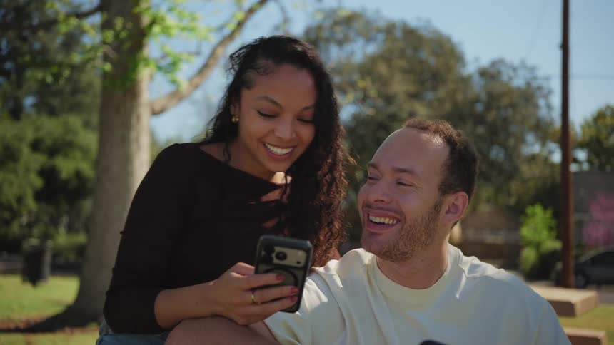 A happy young couple sits together in a park, smiling and laughing while looking at a smartphone on a sunny day