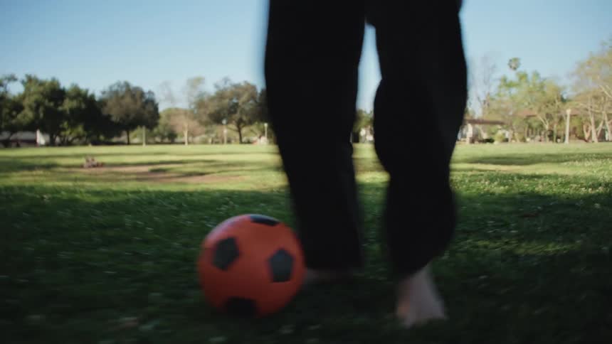 An energetic point-of-view shot follows a soccer ball as people play barefoot on a sunny day in the park