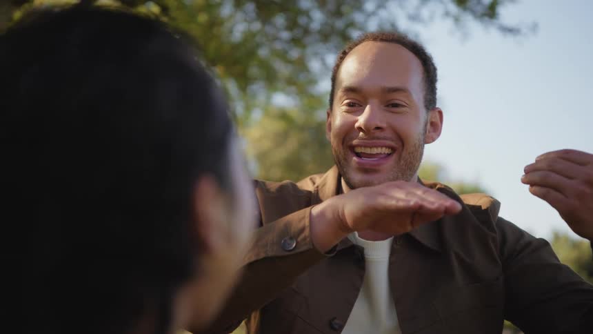 A happy man excitedly recounts a story to a friend, using animated hand gestures in a sunlit park
