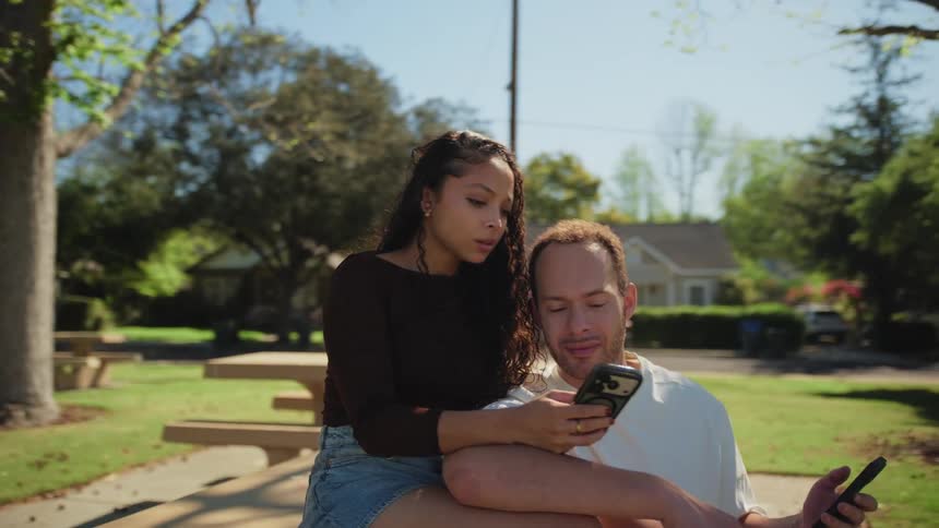 A young, diverse couple sits close together on a picnic table in a sunny park, sharing a moment while scrolling on a smartphone