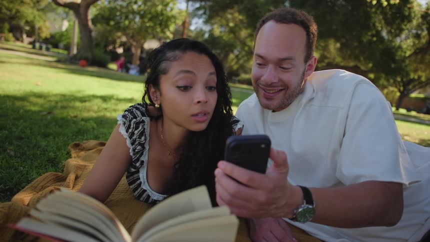 A young couple laughs while looking at a smartphone together during a relaxing picnic in a sunny park