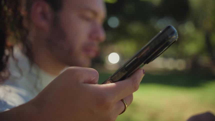 A slow-motion close-up of a hand with painted fingernails scrolling on a smartphone, with a man out of focus in a sunny park