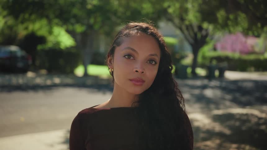 A pensive young woman with long curly hair closes her eyes while standing on a sunny suburban street