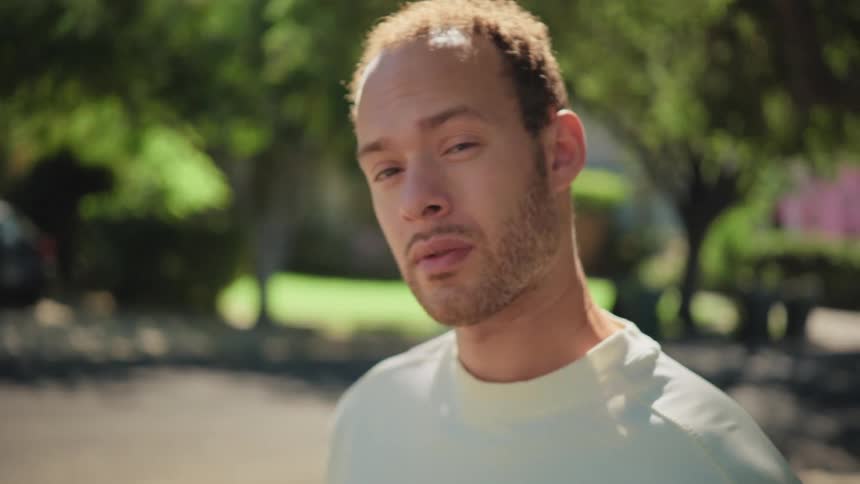 A thoughtful young man makes direct eye contact with the camera while standing in a sunny neighborhood