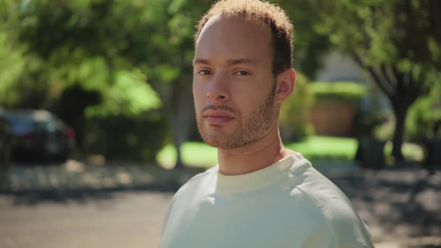 A young man stands outdoors on a sunny day in a suburban neighborhood looking directly at the camera