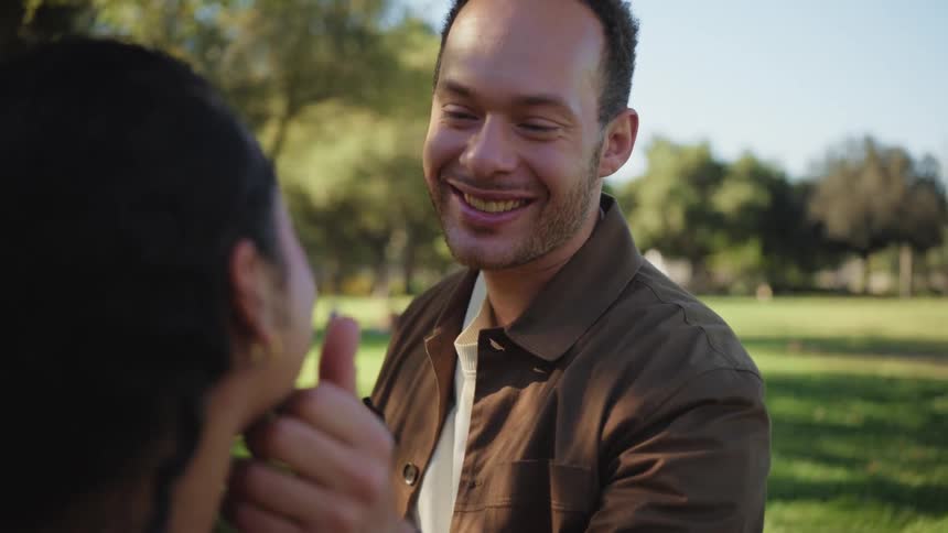 A man lovingly smiles and caresses his partner's face during a romantic moment in a sunny park