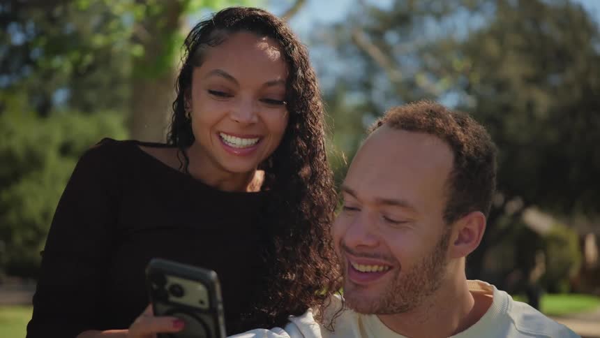A young mixed-race couple smiles and laughs together while looking at a smartphone in a sunlit park