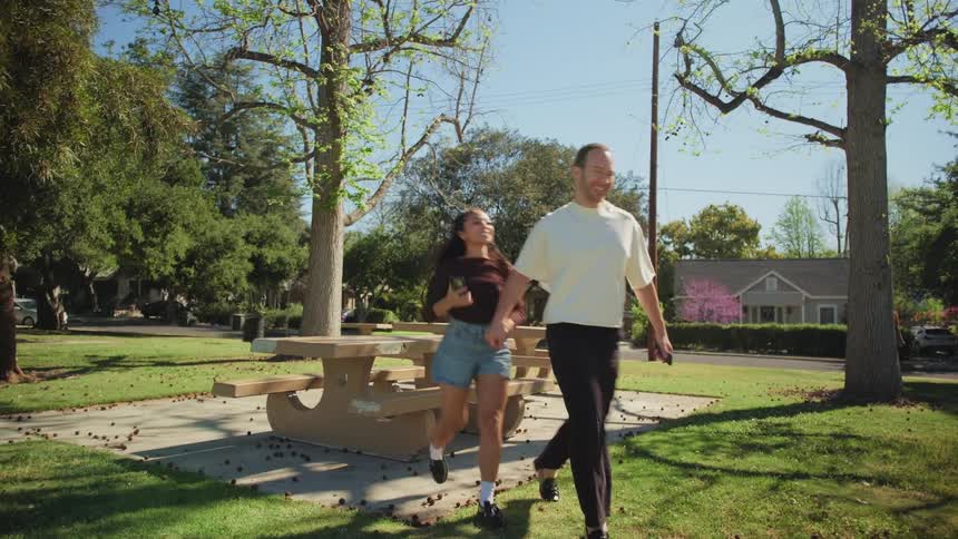 A happy young couple talks and laughs on a picnic table in a sunny park before holding hands and running off together